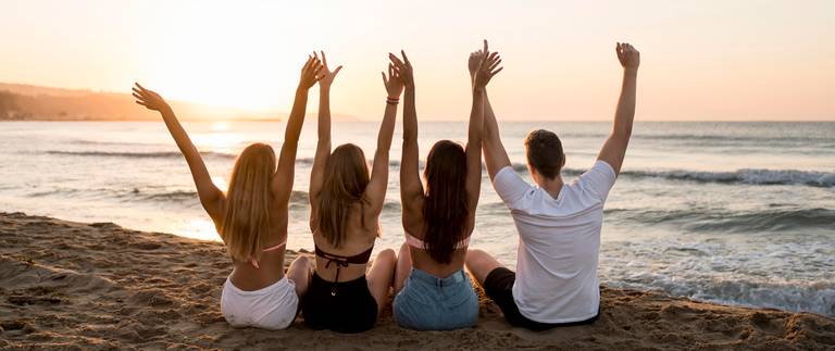 Gruppe von Freunden, die am Meer sitzen und die Arme in die Luft strecken