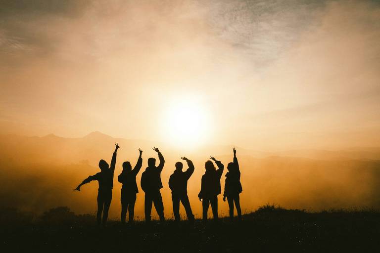 Sechs Personen als Silhouetten, die gemeinsam auf einem Berggipfel stehen – Gegenlicht bei Sonnenaufgang oder -untergang.